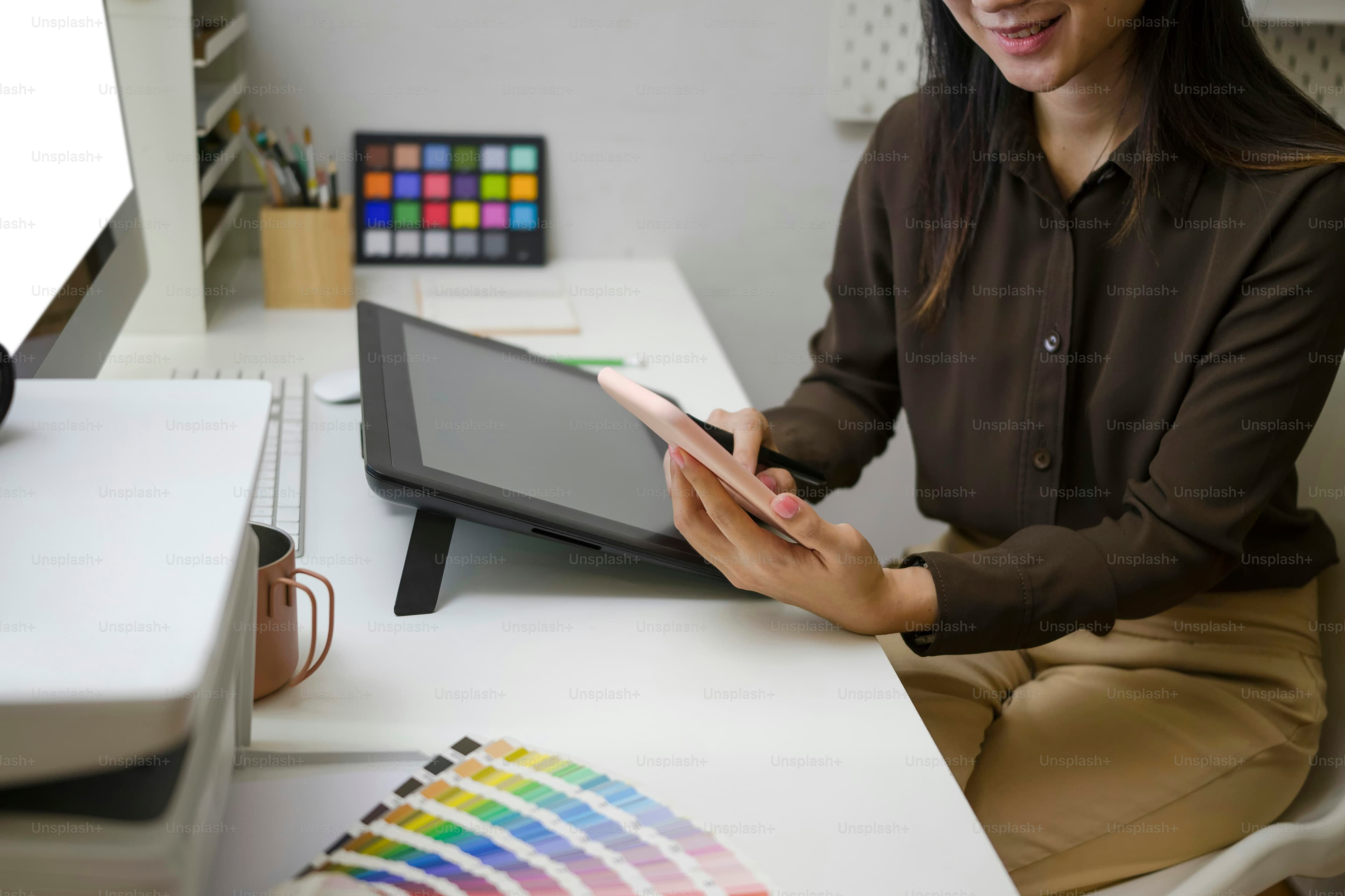 Cropped shot of smiling female graphic designer sitting at her workplace and using mobile phone for texting or massaging with her friends.